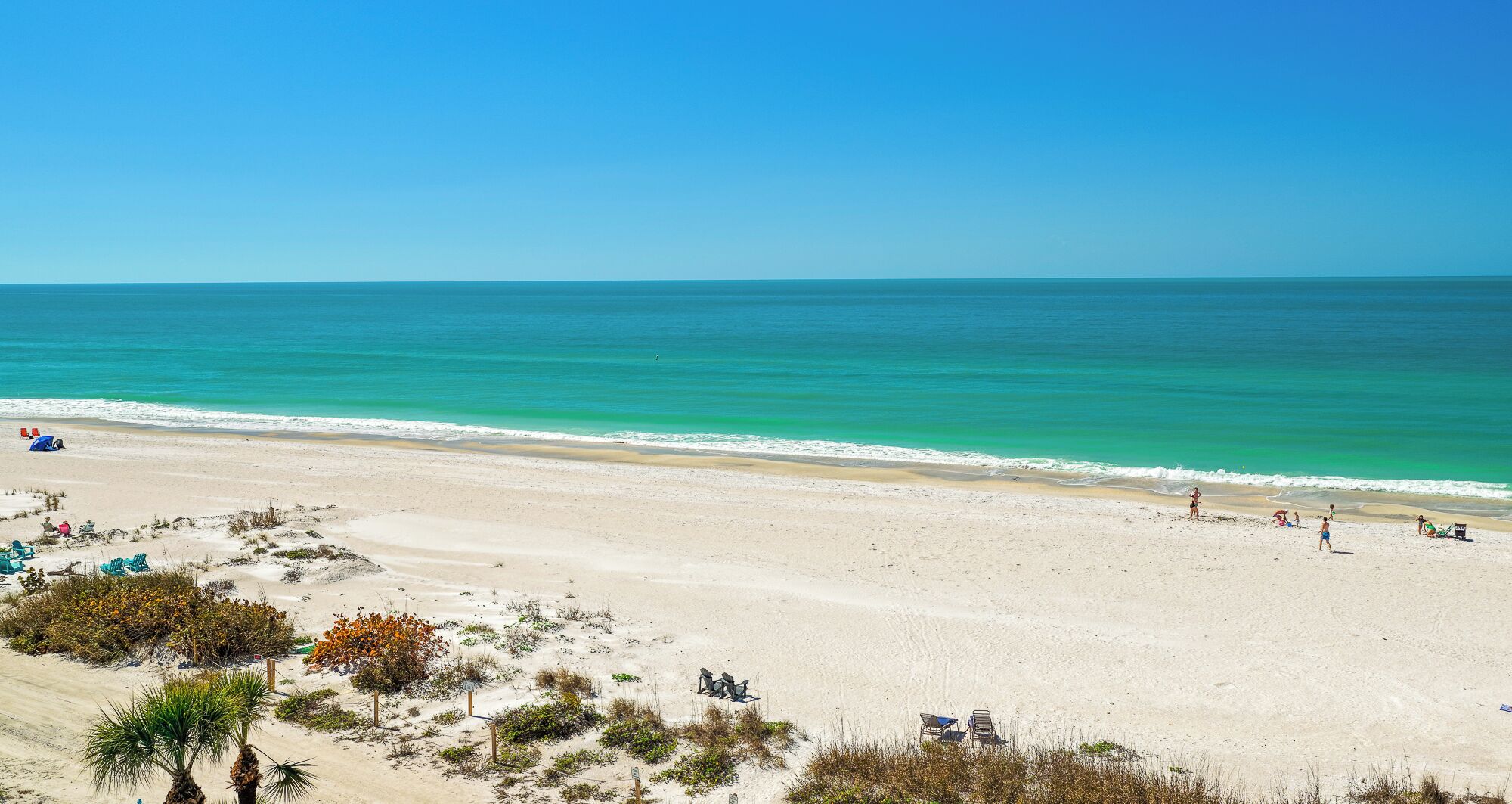 A wide sandy beach with clear turquoise water, a few people, and some beach chairs and umbrellas.