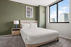 Habitación de hotel con cama, mesita de noche, lámpara, un cuadro en la pared y ventana con vista a la ciudad.