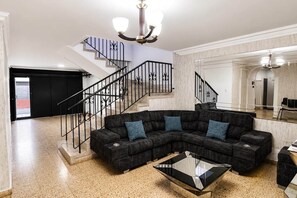 A spacious living room with a black sectional sofa, a glass coffee table, a staircase with black railings, and a large mirror reflecting the room.