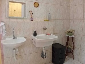 Ein Badezimmer mit zwei Waschbecken, einem Spiegel, einem Handtuchhalter, einem Ablagebereich mit Toilettenartikeln und einem kleinen hölzernen Hocker mit einer Pflanze.
