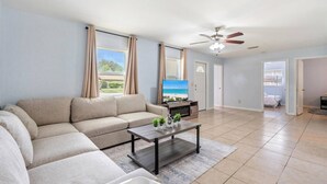 A living room with a beige sofa, a coffee table, and a TV. There is a ceiling fan and a view of the outdoors through the windows. - King-Style Retreat · 4 BR · Family Friendly (Tampa)