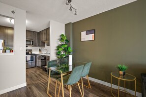 Una cocina moderna con electrodomésticos de acero inoxidable, un comedor con mesa de vidrio y sillas verdes, y una pequeña mesa redonda con una planta.