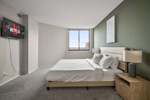 Un dormitorio moderno con una cama grande, un televisor montado en la pared, una ventana y una mesita de noche con una lámpara.