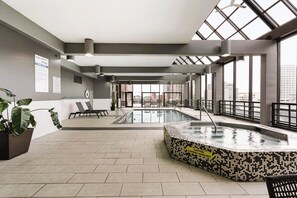Un área de piscina interior moderna con un jacuzzi, sillas de descanso y amplias ventanas con vistas a la ciudad.