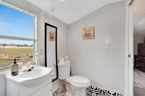 Une salle de bain avec un lavabo, des toilettes et un miroir, tous de couleur blanche. Deux tableaux encadrés sont accrochés au mur.