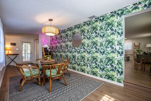 Un coin repas avec une table ronde et des chaises, un tapis à motifs et un mur orné d’un motif de feuilles tropicales.