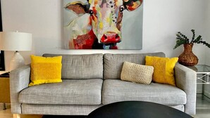 Un soggiorno con un divano grigio, cuscini gialli, un tavolino da caffè nero, una lampada e un quadro di una mucca con il testo "Get in the Texas mood!".