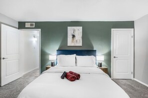 A hotel room with a bed, two bedside tables with lamps, a painting on the wall, and two doors.