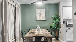 A dining area with a wooden table, chairs, a ceiling fan, a wall-mounted painting, and a potted plant.