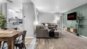 A modern living room with a grey sofa, a dining area with a wooden table and chairs, a kitchen in the background, and a TV mounted on the wall displaying 'NETFLIX'.