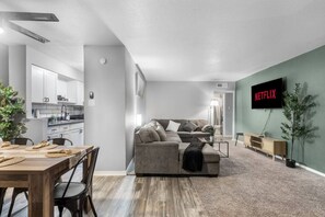 A modern living room with a grey sofa, a dining area with a wooden table and chairs, a kitchen in the background, and a TV mounted on the wall displaying 'NETFLIX'.