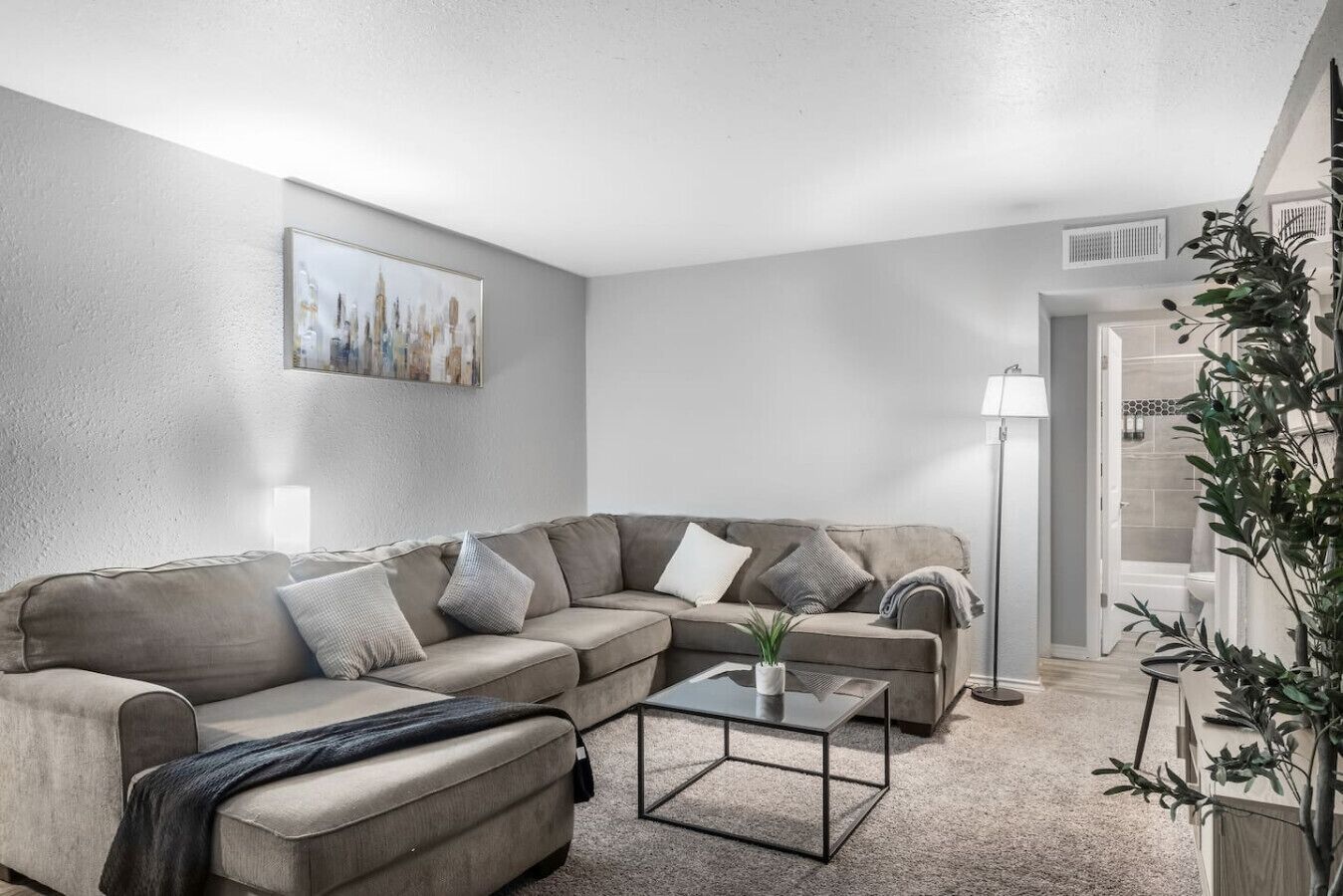 Ein modernes Wohnzimmer mit einem beigefarbenen Ecksofa, einem Glastisch und einem Stadtbild an der Wand.