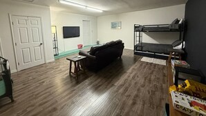 Un soggiorno con pavimento in legno marrone scuro, un divano in pelle nera, un tavolino in legno e una cabina letto, con un televisore appeso al muro.