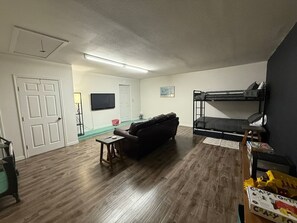 Ein Wohnzimmer mit dunklem Holzboden, einer schwarzen Ledersofa, einem kleinen Holztisch, einem Etagenbett und einem Fernseher an der Wand.