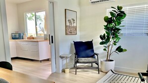 Un salon moderne avec une table à manger en bois, une chaise bleue, une plante en pot et une vue sur la cuisine par une porte ouverte.