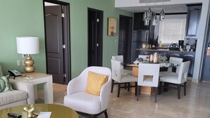Ein modernes Wohnzimmer mit Sofa, Sessel und Couchtisch, ein Essbereich mit Stühlen und Tisch sowie eine Küche mit Geräten und einem Fenster.
