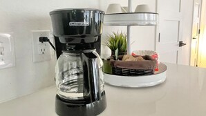 Eine Kaffeemaschine steht auf einer weißen Arbeitsplatte, im Hintergrund eine weiße Wand und eine Tür.