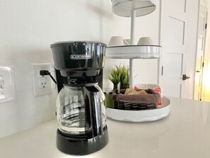Eine Kaffeemaschine steht auf einer weißen Arbeitsplatte, im Hintergrund eine weiße Wand und eine Tür.