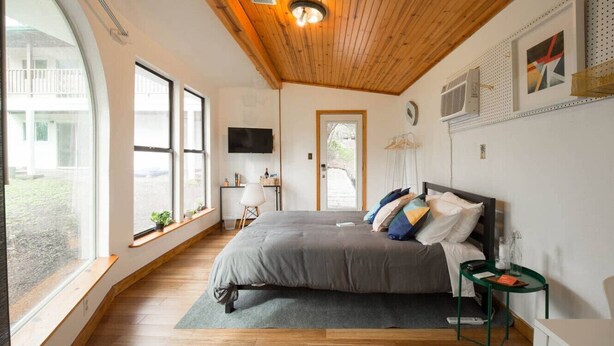 A bedroom with a large bed, wooden flooring, a ceiling fan, a TV, and a window with a view of the outside. - East Austin Garden Cottage |  Sweet & Private (Austin)