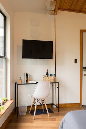 A modern room with a flat-screen TV mounted on the wall, a small desk with a chair, a wicker basket, and a potted plant.