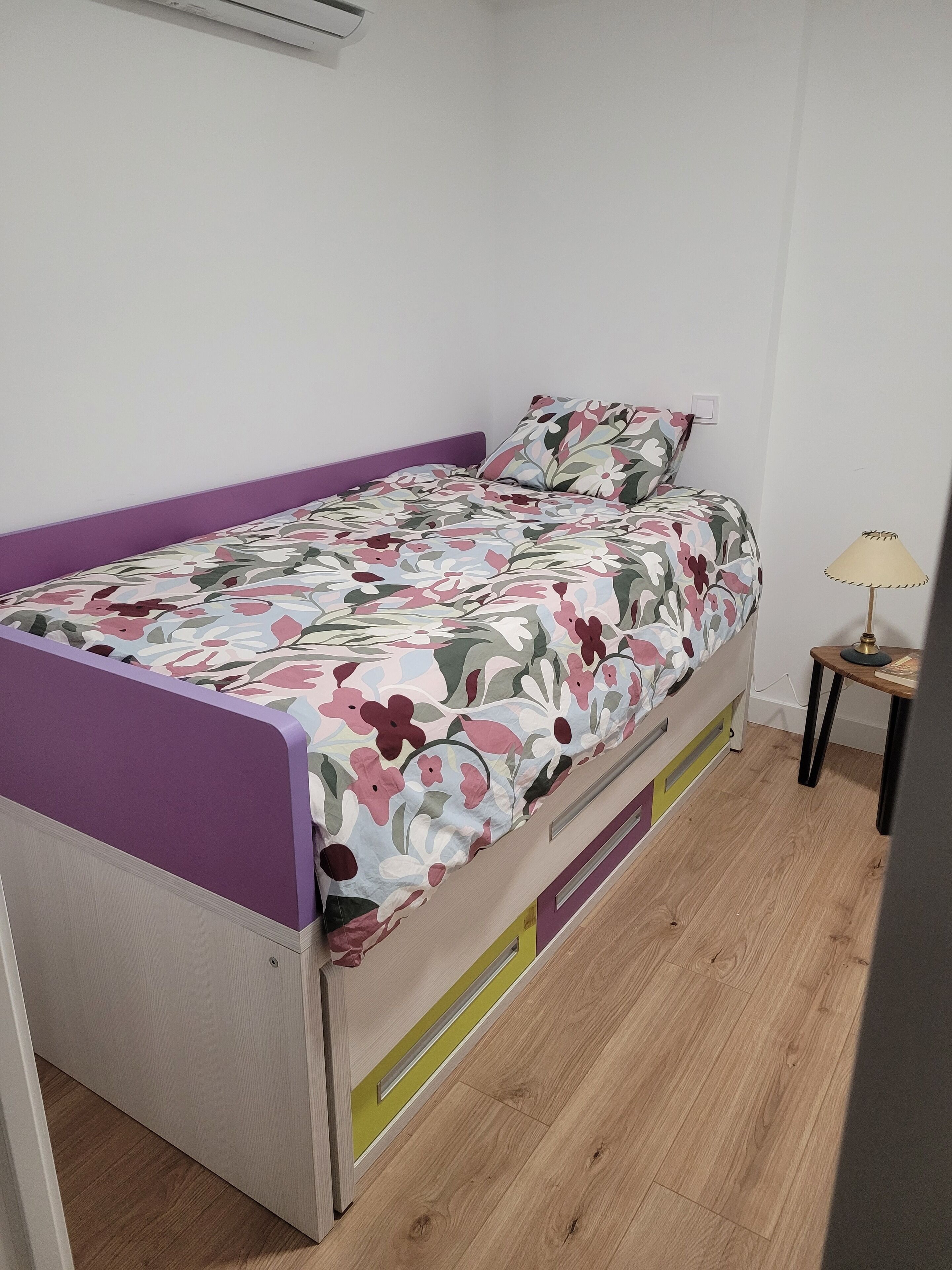 A single bed with a floral bedspread, a purple headboard, and a small bedside table with a lamp.