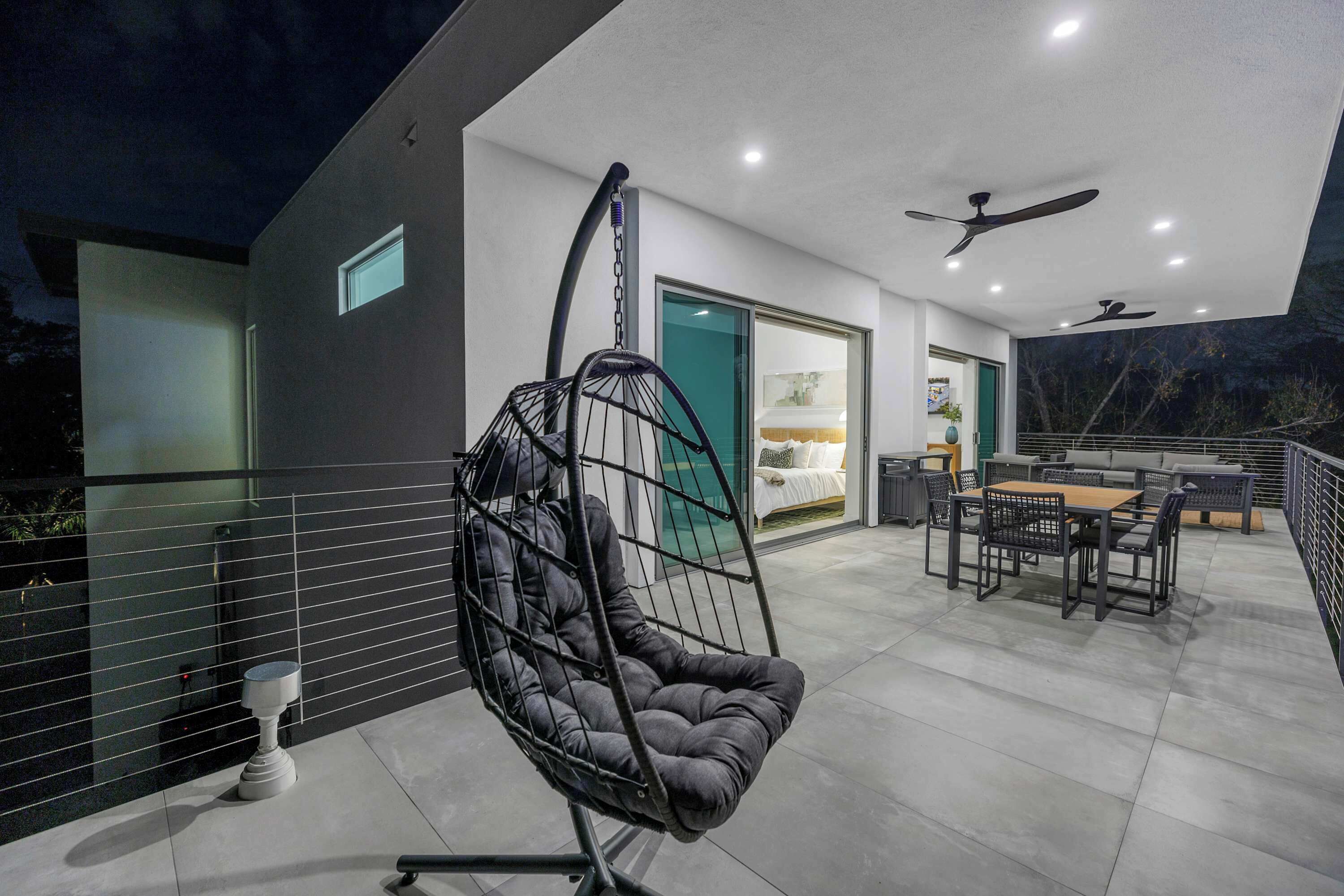 A modern outdoor patio with a hanging chair, a dining table with chairs, and a ceiling fan.
