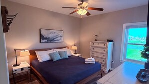 A bedroom with a bed, nightstands, a dresser, a ceiling fan, a window with a view, and a painting on the wall. - Bolivar Beachside Getaway (Crystal Beach)