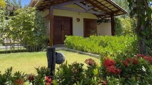 A single-story building with a sloped roof, surrounded by lush greenery and flowering plants. - Pousada Rancho Paraiso (Porto de Pedras)