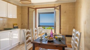 Eine Küche mit weißen Schränken, einem Esstisch mit Stühlen, Blick aufs Meer durch eine offene Tür und eine kleine Kücheninsel mit Spüle und Toaster.