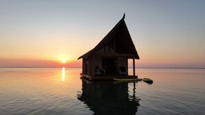 A thatched-roof bungalow on stilts over calm water, with a sunset in the background. - The Grand Tiki at Tiki Suites – A Floating Oasis in Key West (Key West)