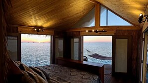 A wooden cabin with a bed, a small table, and a hammock. It has large windows overlooking the sea. - The Grand Tiki at Tiki Suites – A Floating Oasis in Key West (Key West)