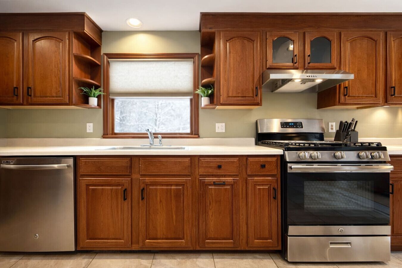 A modern kitchen with wooden cabinets, stainless steel appliances, and a window with a white blind.