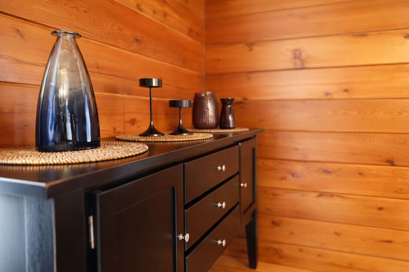 Una habitación con paneles de madera, un aparador de madera oscura, un florero de vidrio transparente y dos lámparas negras sobre caminos de mesa tejidos.