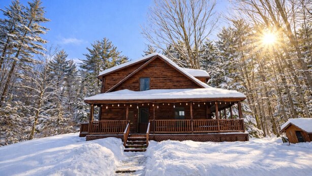 Une cabane en bois dotée d’un porche et d’un escalier, entourée d’arbres couverts de neige et sous un ciel dégagé.