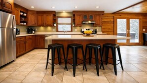 Une cuisine moderne avec des meubles en bois, un réfrigérateur en acier inoxydable, un grand îlot central doté de tabourets de bar et une fenêtre donnant sur un paysage enneigé.