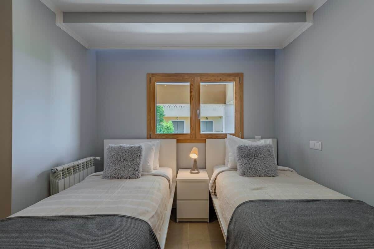 A bedroom with two beds, a window with a view, a radiator, and a nightstand with a lamp.