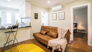 En kompakt opholdsstue med tekøkken, sofa, spiseplads og et lille bord med lampe.