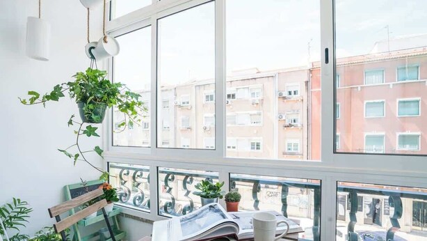 En balkon med en træbænk, et bord med et magasin og en kop, krukker med planter og udsigt til lejlighedskomplekser.