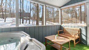 Un bain à remous doté d’un banc et d’une table en bois, entouré d’une zone abritée avec des fenêtres donnant sur un paysage enneigé.