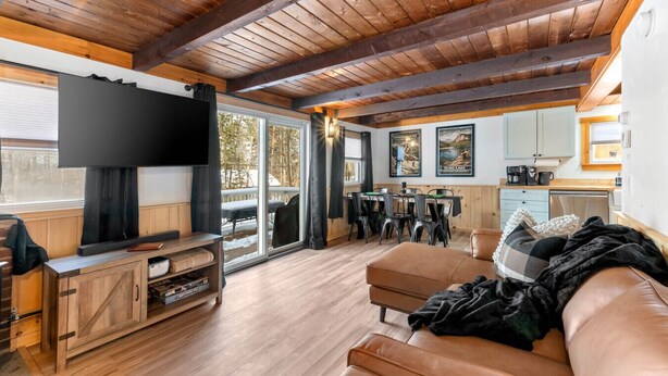 Un salon chaleureux doté d’un grand téléviseur à écran plat, de poutres apparentes en bois au plafond, d’un canapé en cuir et d’un coin repas avec une table et des chaises.