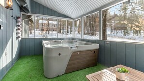 Un bain à remous situé dans un espace extérieur abrité, avec du gazon synthétique, une table en bois et une vue sur un paysage enneigé.