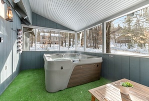 A hot tub in a covered outdoor area with artificial grass, a wooden table, and a view of a snowy landscape.