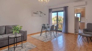 A modern living room with a grey sofa, a dining table with chairs, a chandelier, and a large window with curtains. - Casa Rocha Portimao with Terrace (Portimão)