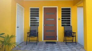 A yellow building with a brown door, two chairs with patterned cushions, and a potted plant. - Central & Secure Stay - Minutes from Town, Airport and Stadium (Osbourn)