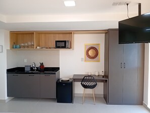 Cozinha compacta com armários cinzentos, bancada preta, micro-ondas, pia e um cesto de lixo preto. Há uma pequena área de refeições com uma cadeira e uma mesa, e uma TV fixada na parede.