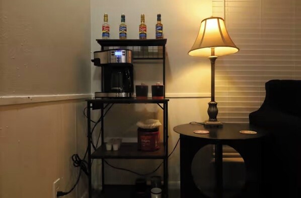 Een kleine kamer met een zwarte plank waarop flessen, een koffiezetapparaat en een lamp staan.