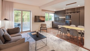 A modern living room with a sofa, coffee table, and a TV. There is a dining area with a table and chairs, and a kitchen with appliances. - Premium 2BR Apartment in Avenidas Novas, Lisbon | Balcony & Included Services (Lisboa)
