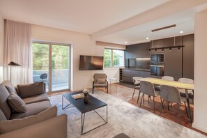 A modern living room with a sofa, coffee table, and a TV. There is a dining area with a table and chairs, and a kitchen with appliances. - Premium 2BR Apartment in Avenidas Novas, Lisbon | Balcony & Included Services (Lisboa)