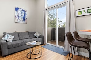 Ein Wohnzimmer mit grauem Sofa, einem kleinen runden Tisch, zwei braunen Sesseln, einer Schiebeglastür und einem gerahmten Bild an der Wand.