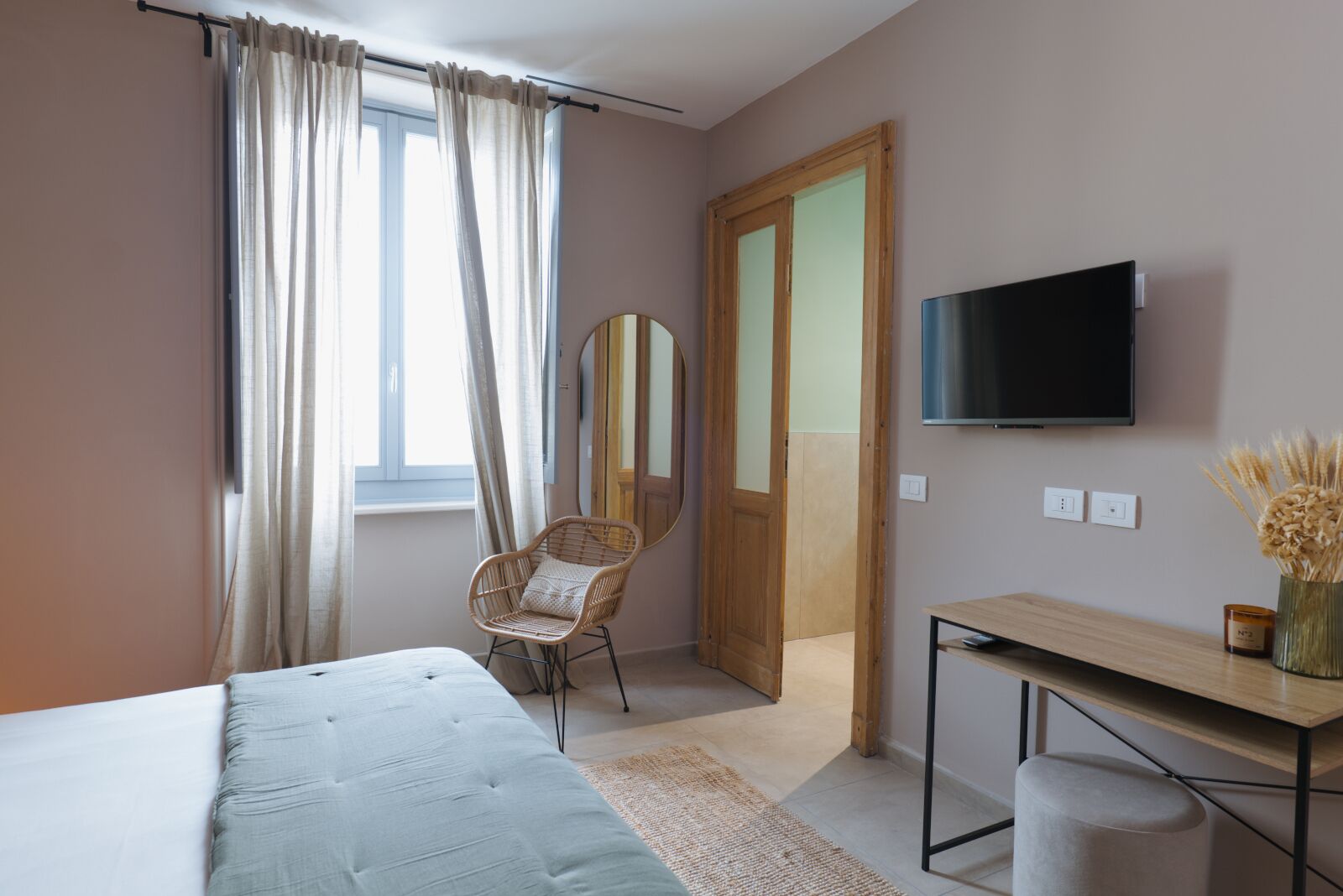 Un dormitorio con cama, una silla, televisor, escritorio y una puerta que da a otra habitación.